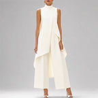 A model wearing a sleeveless, high-neck white jumpsuit with a dramatic draped overlay and wide-leg pants, styled with white high-heeled sandals against a neutral gray studio background.