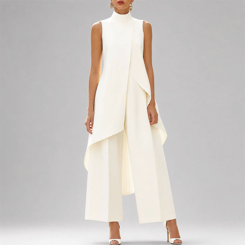 A model wearing a sleeveless, high-neck white jumpsuit with a dramatic draped overlay and wide-leg pants, styled with white high-heeled sandals against a neutral gray studio background.