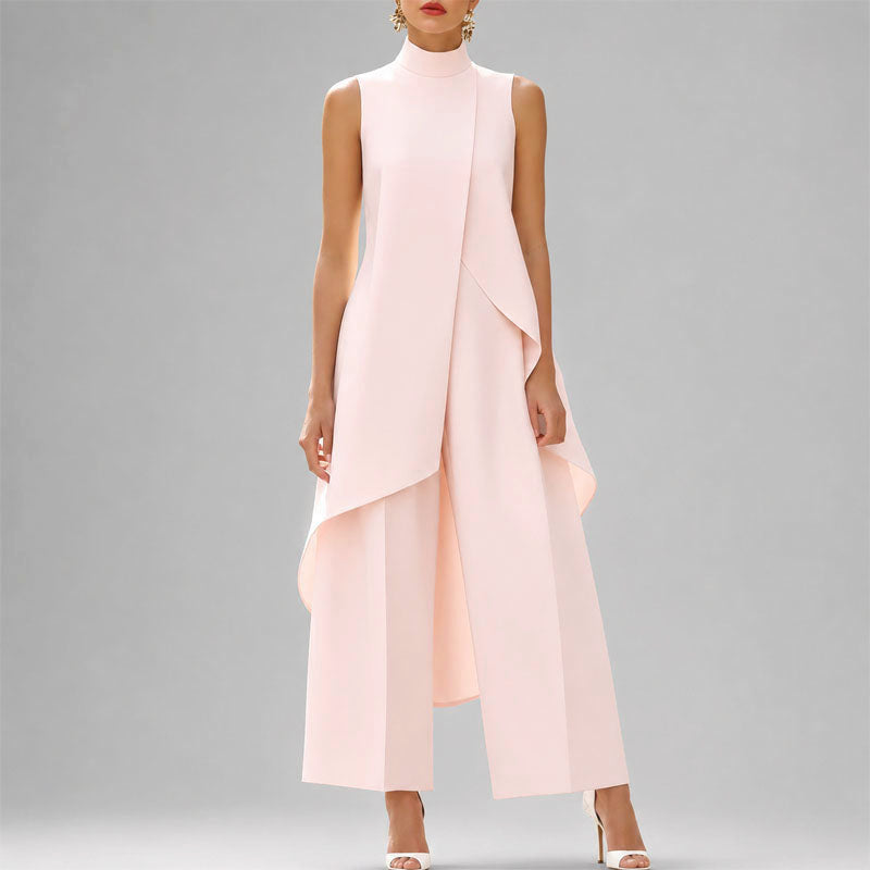 A model wearing a sleeveless, high-neck, light pink jumpsuit with an asymmetrical draped overlay and wide-leg cropped pants, set against a solid gray studio background.