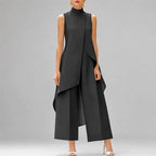 A model wearing a sleeveless black high-neck jumpsuit with draped asymmetrical layers and wide-leg cropped pants, paired with white high-heeled sandals against a solid gray studio background.