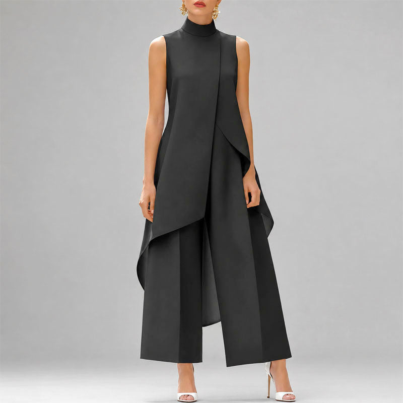 A model wearing a sleeveless black high-neck jumpsuit with draped asymmetrical layers and wide-leg cropped pants, paired with white high-heeled sandals against a solid gray studio background.